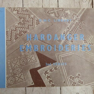 Könnte beinhalten: Ein Vintage-Buchcover mit einem blau-weiß gestreiften Rand. Der Titel lautet "Hardanger Embroideries" mit dem Untertitel "2nd Series". Das Cover zeigt verschiedene weiße Stickmuster auf braunem Hintergrund.