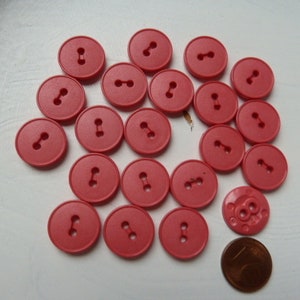 May include: A collection of 18 pink plastic buttons with two holes each. The buttons are arranged in a circle on a white surface.