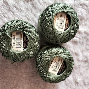 Könnte beinhalten: Drei Knäuel olivgrünes Perlgarn. Jedes Knäuel hat ein Etikett mit dem Text "Col. 860" und "Pearl Cotton 8". Der Faden ist eng gewickelt und erzeugt eine strukturierte Oberfläche.