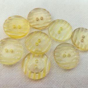 Könnte beinhalten: Acht durchscheinende gelbe Knöpfe mit jeweils zwei oder drei Löchern. Die Knöpfe sind rund und haben ein gestreiftes Design. Sie liegen auf einem weißen Stoff. Die Knöpfe sind hellgelb.