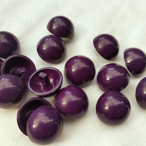 Pode incluir: Uma coleção de botões roxos escuros e brilhantes espalhados sobre uma superfície branca. Os botões são redondos com uma forma ligeiramente abaulada e uma haste na parte de trás. Alguns botões estão virados para cima, outros mostram a haste.