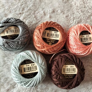 May include: Five spools of Anchor Pearl Cotton 8 Coton Perle thread in various colors: gray, copper, coral, light blue, and brown. Each spool has a label with a color code and barcode. The thread is tightly wound into a ball shape.