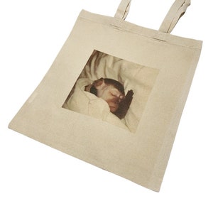 May include: A beige tote bag featuring a printed image of a baby monkey sleeping in a white blanket.