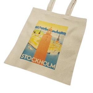 May include: A beige canvas tote bag with a vintage-style illustration of the Stockholm skyline. The illustration features a tall red tower, buildings, and boats on a blue body of water. The text "STOCKHOLM" is printed in black at the bottom of the illustration.