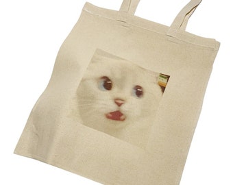 Op de afbeelding: Een witte canvas tote bag met een bedrukte afbeelding van een witte kat met blauwe ogen die er verbaasd uitziet. De mond van de kat is open en zijn tong steekt eruit.