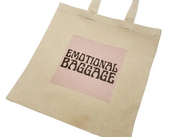 May include: A beige canvas tote bag with pink and brown text that reads "Emotional Baggage".