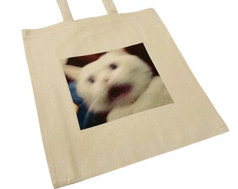 May include: A white tote bag with a printed image of a white cat with wide eyes and an open mouth. The cat appears to be startled or surprised.