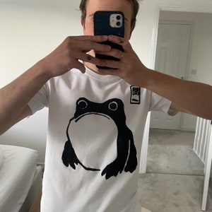 May include: White t-shirt featuring a large black frog graphic on the front. The frog has a simple, bold design. A small square graphic with Japanese text is on the upper right. The shirt is short-sleeved.