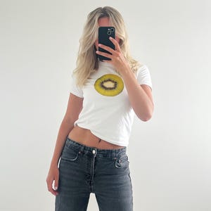 May include: A white t-shirt with a graphic of a kiwi fruit slice. The shirt is worn by a person with blonde hair. The person is holding a black smartphone in front of their face.
