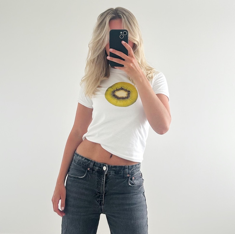May include: A white t-shirt with a graphic of a kiwi fruit slice. The shirt is worn by a person with blonde hair. The person is holding a black smartphone in front of their face.