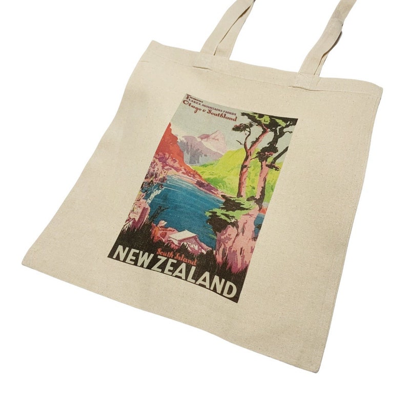 Peut inclure: Sac fourre-tout de couleur cr&egrave;me avec un design d'affiche de voyage vintage. L'affiche repr&eacute;sente un paysage pittoresque avec des montagnes, de l'eau et des arbres, avec les mots "NEW ZEALAND" affich&eacute;s.