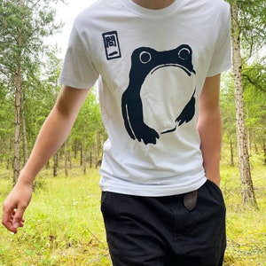 May include: White t-shirt featuring a large black frog graphic. The shirt also has Japanese text in a black box on the upper left. The person is wearing black pants and is standing in a forest.