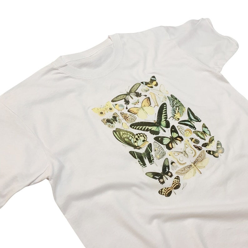 以下が含まれることがあります： 白のTシャツ。前面には、緑、黄色、黒など様々な色の蝶のイラストが描かれています。蝶は長方形の形に配置されています。Tシャツはクルーネックで半袖です。