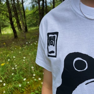 May include: White t-shirt with a black graphic design. The design features a large black shape with a white circle and a smaller black rectangular design with Japanese characters. The shirt is made of a soft material.