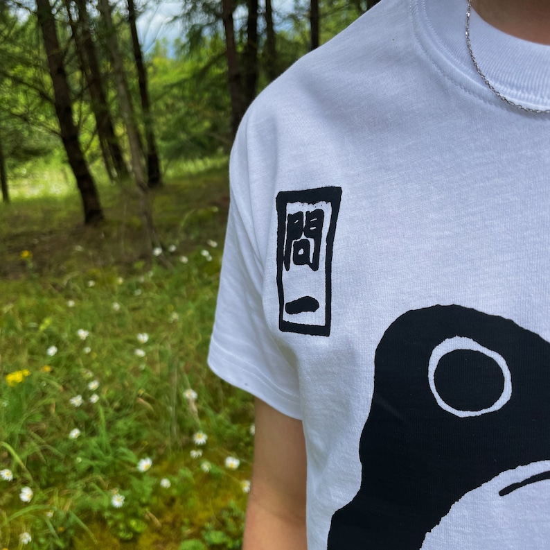 May include: White t-shirt with a black graphic design. The design features a large black shape with a white circle and a smaller black rectangular design with Japanese characters. The shirt is made of a soft material.