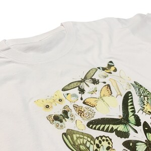以下が含まれることがあります： 白のTシャツ。前面には、黄色、緑、黒など様々な色の蝶の繊細なイラストが描かれています。蝶はTシャツの前面に集まって配置されています。