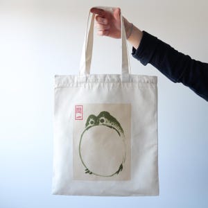 May include: A white canvas tote bag with a green frog illustration and a red Japanese character. The frog is depicted in a circle shape with its head and legs visible.