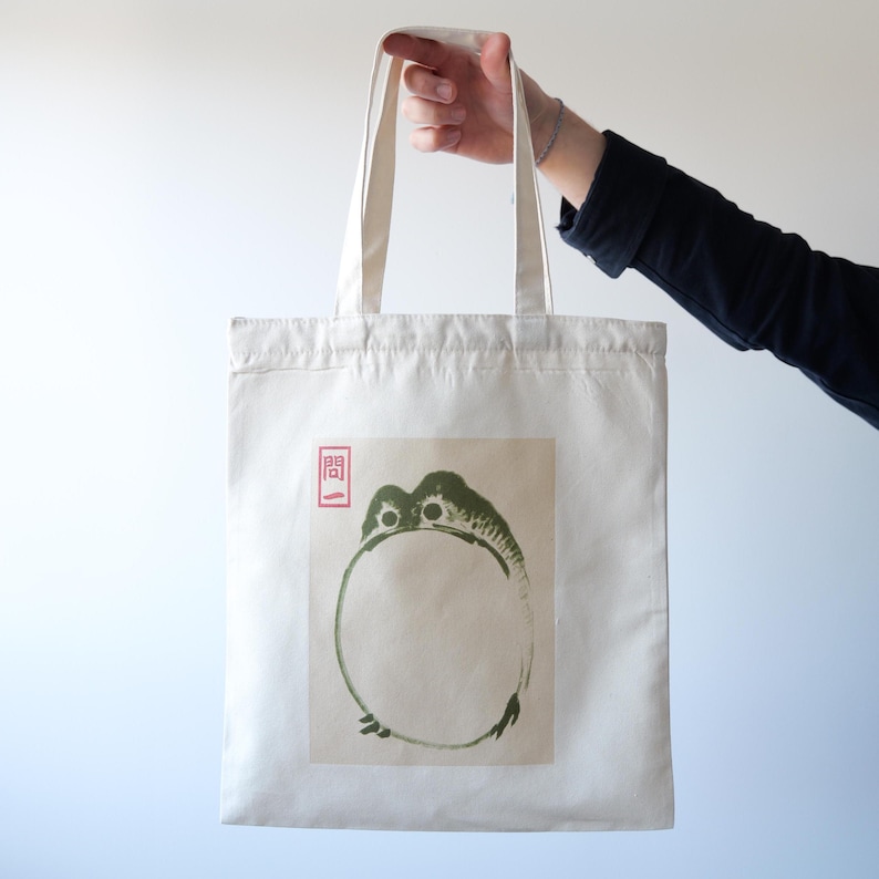 May include: A white canvas tote bag with a green frog illustration and a red Japanese character. The frog is depicted in a circle shape with its head and legs visible.