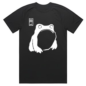 May include: Black t-shirt featuring a white graphic of a frog with a large open mouth. Above the frog is a small white square with a black symbol. The shirt is a casual wear item.