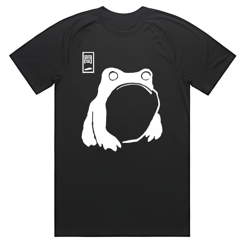 May include: Black t-shirt featuring a white graphic of a frog with a large open mouth. Above the frog is a small white square with a black symbol. The shirt is a casual wear item.