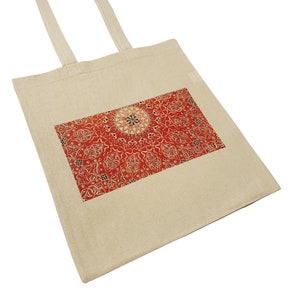May include: A beige canvas tote bag with a red and white geometric pattern printed on the front. The pattern features a central circle with intricate details and radiating lines.