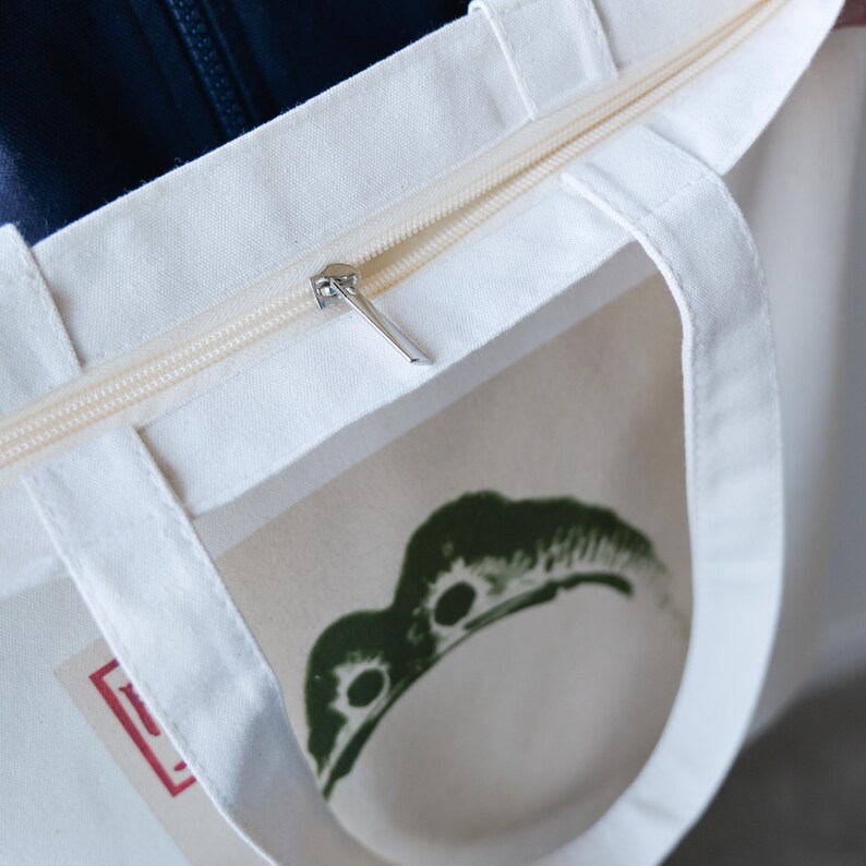 May include: A white canvas tote bag with a zipper closure and a green graphic print. The bag has two handles and a zippered pocket on the front.