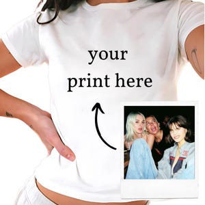 Könnte beinhalten: Weißes T-Shirt mit dem Text "your print here" und einem Pfeil, der auf die Mitte des Shirts zeigt. Neben dem Text ist ein Polaroid-Foto von drei Personen zu sehen.