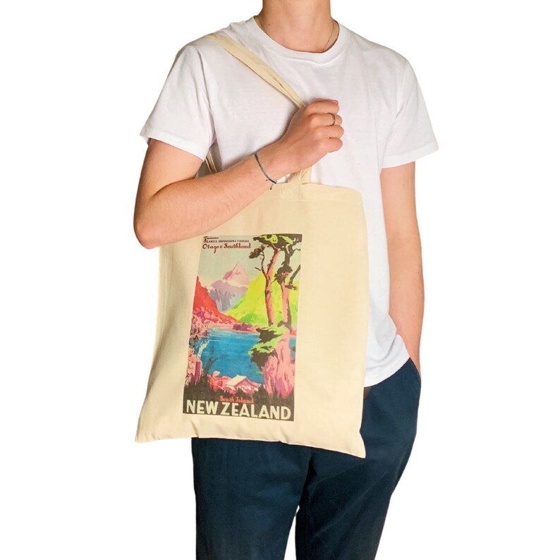 Peut inclure: Sac fourre-tout cr&egrave;me avec un design d'affiche de voyage vintage pour la Nouvelle-Z&eacute;lande. L'affiche repr&eacute;sente un paysage pittoresque avec des montagnes, un lac et des arbres, avec les mots "NEW ZEALAND" en &eacute;vidence. Le sac a une forme simple et rectangulaire avec de longues sangles.