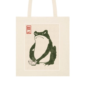 Può includere: Una borsa tote con un'illustrazione di una rana verde in stile giapponese. La rana tiene una ciotola e un cucchiaio di legno. La parola "開" è scritta in rosso sulla borsa.