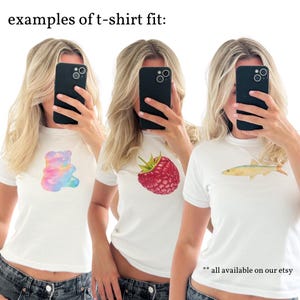 May include: Three women model white t-shirts with different graphic designs. The designs include a rainbow gummy bear, a red raspberry, and a yellow fish. The text "**all available on our etsy" is at the bottom of the image.