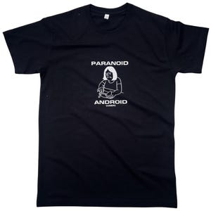 Puede incluir: Camiseta negra con un gráfico y texto en blanco. El gráfico representa a una persona con expresión triste usando un dispositivo. El texto dice "PARANOID ANDROID (USER)".