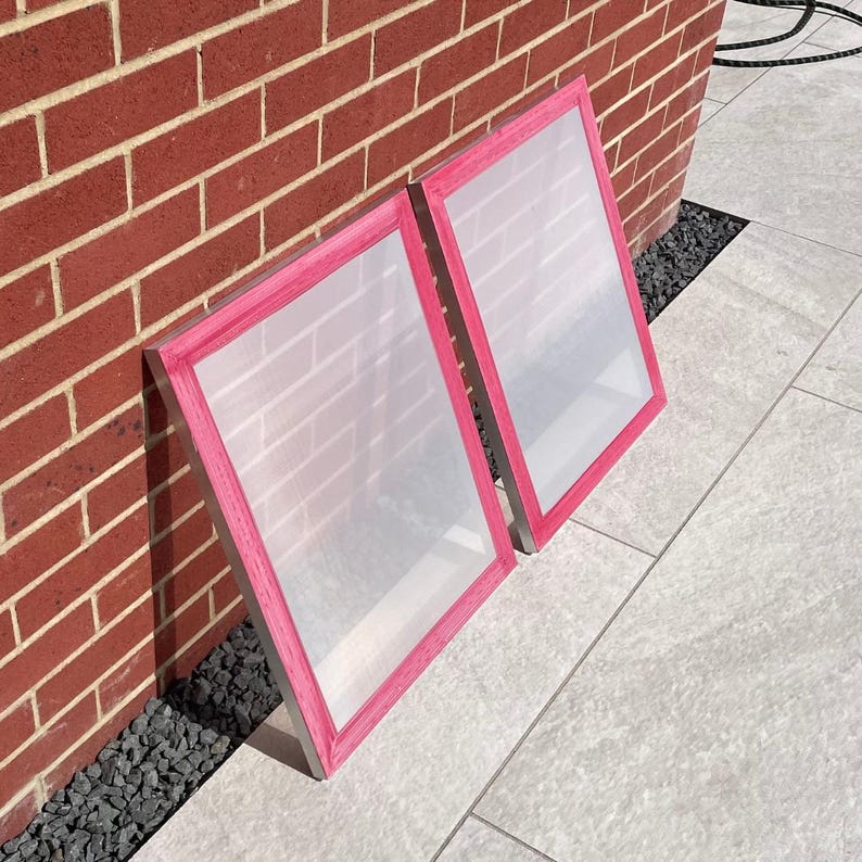 May include: Two screen printing frames with pink tape around the edges. The frames are made of metal and have a mesh screen stretched across them.