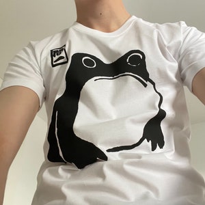May include: White t-shirt featuring a large black frog graphic. The frog design is centered on the shirt and has a minimalist style. A small square graphic is on the upper left. The shirt is made of a soft material.