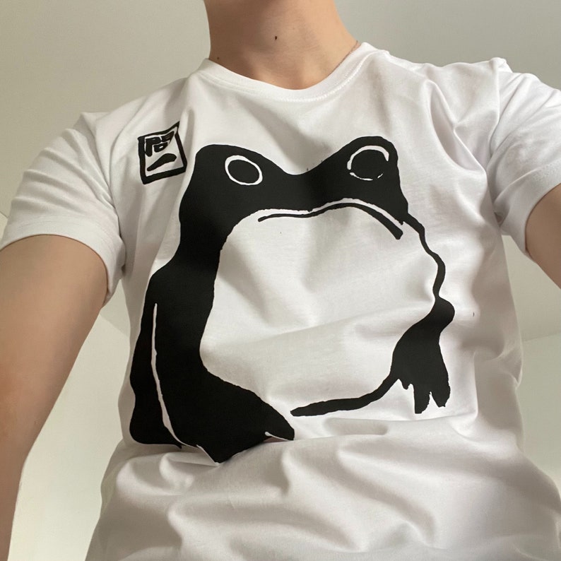 May include: White t-shirt featuring a large black frog graphic. The frog design is centered on the shirt and has a minimalist style. A small square graphic is on the upper left. The shirt is made of a soft material.