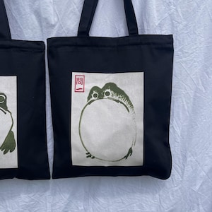 May include: Black tote bag with a white square design featuring a green frog with the Japanese character "開" in red.
