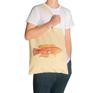May include: A beige tote bag with a red and yellow fish illustration. The bag has a long strap and is being held by a person.