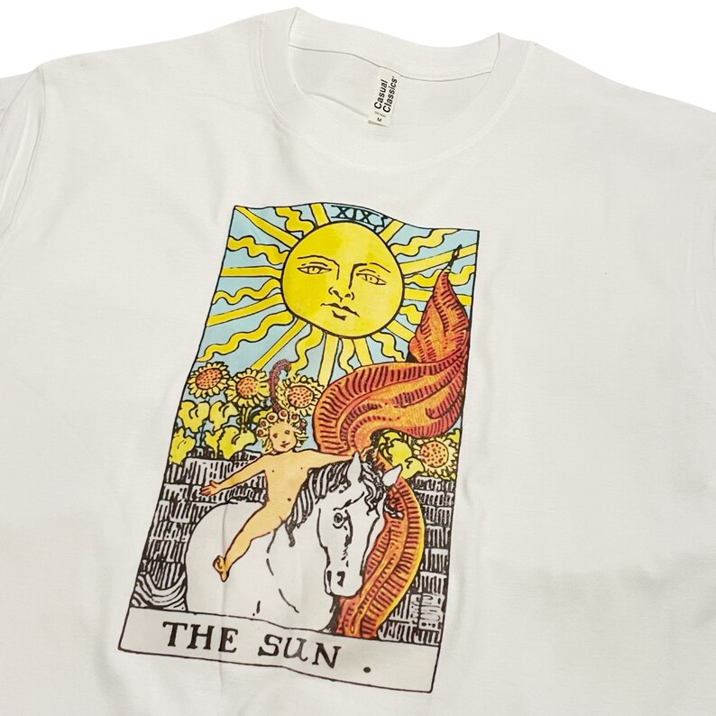 May include: White t-shirt featuring a colourful tarot card design of the Sun card. The card shows a sun with a face, a child riding a white horse, and sunflowers.