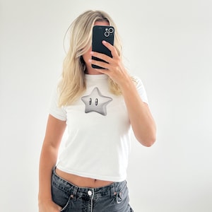 May include: White cropped t-shirt with a silver star graphic. The star has two black eyes.