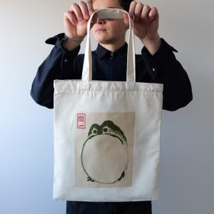 May include: A white canvas tote bag with a green and black frog illustration and the word "BAI" in red. The frog is holding a circle in its hands.