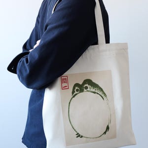 May include: A white canvas tote bag with a green and black ink illustration of a frog on the front. The frog is in a circle and the word "開" is in red ink above the frog.