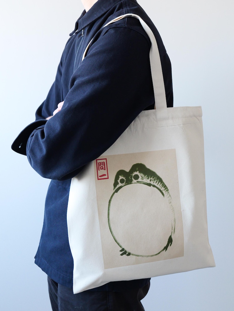 May include: A white canvas tote bag with a green and black ink illustration of a frog on the front. The frog is in a circle and the word "開" is in red ink above the frog.