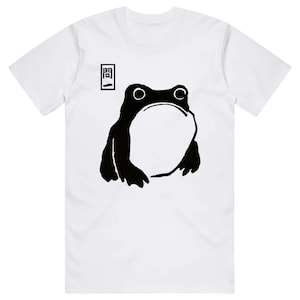 May include: White t-shirt featuring a black illustration of a frog with a large open mouth. The frog design is minimalist and bold. Above the frog is a small rectangular graphic with Japanese text. The shirt has short sleeves.