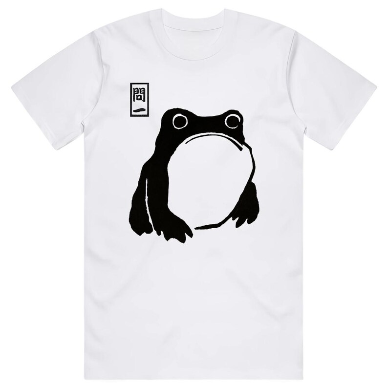 May include: White t-shirt featuring a black illustration of a frog with a large open mouth. The frog design is minimalist and bold. Above the frog is a small rectangular graphic with Japanese text. The shirt has short sleeves.