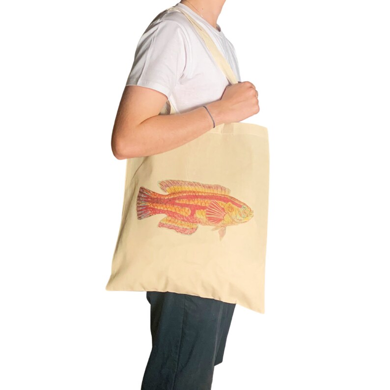May include: A person is holding a beige tote bag with a red and yellow fish illustration. The bag has a long strap and is made of canvas.