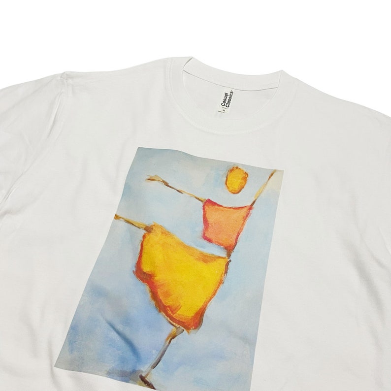 Op de afbeelding: Wit T-shirt met een grafische print van een gestileerde persoon in een gele en oranje jurk. Het label heeft de tekst "Casual Classics".