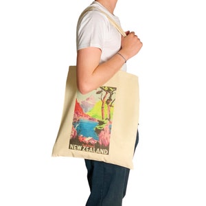 Peut inclure: Sac fourre-tout cr&egrave;me avec une illustration color&eacute;e d'un paysage de Nouvelle-Z&eacute;lande, avec le texte "NEW ZEALAND". Le sac est port&eacute; par une personne. Le paysage comprend des montagnes, de l'eau et des arbres.