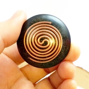 Op de afbeelding: Een ronde, donkergroene steen met een spiraalvormig ontwerp van koperdraad. De steen wordt in een hand gehouden.