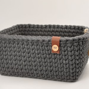 Könnte beinhalten: Ein grauer gehäkelter Aufbewahrungskorb mit einem braunen Ledergriff und einem Holzknopf. Der Korb hat eine gewebte Textur und ist handgefertigt.