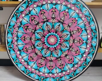 May include: A round, black decorative piece featuring a vibrant mandala design. The mandala showcases a symmetrical pattern with pink, teal, and blue floral elements, accented with white bead-like details. The piece is displayed against a neutral background.