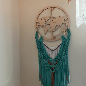 May include: A wooden world map and compass rose wall hanging with a turquoise macrame dream catcher. The dream catcher has white and brown accents, with long fringe. Decorative lights and seashells are in the background.